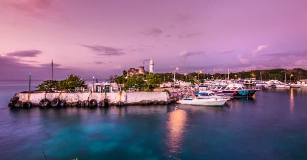Puerto de Cozumel al atardecer con barcos anclados, un faro blanco y cielo en tonos violeta.