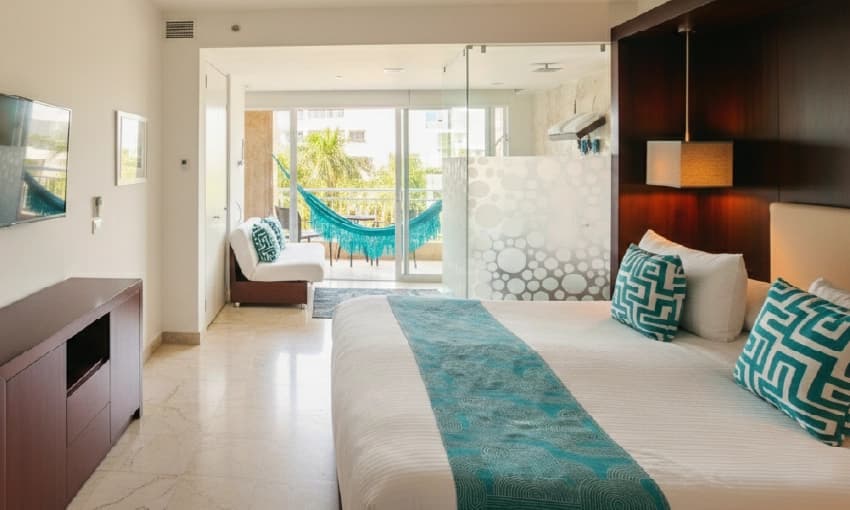 Loft-style room with white bedding, turquoise accents, and terrace overlooking palm trees.