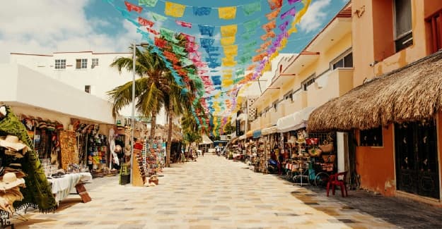 Calle peatonal decorada con banderas coloridas, tiendas de artesanías y palmeras bajo el sol.
