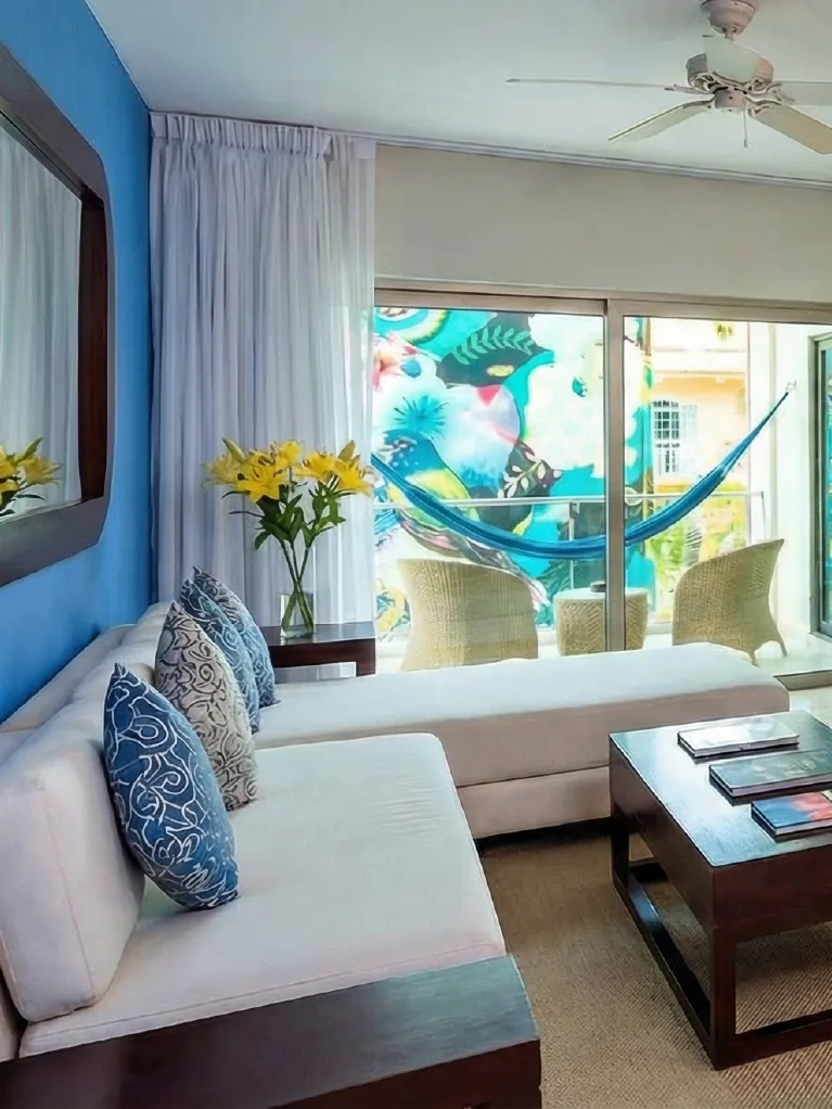 Living area in hotel suite with white sofa, wooden table, and balcony with blue hammock.