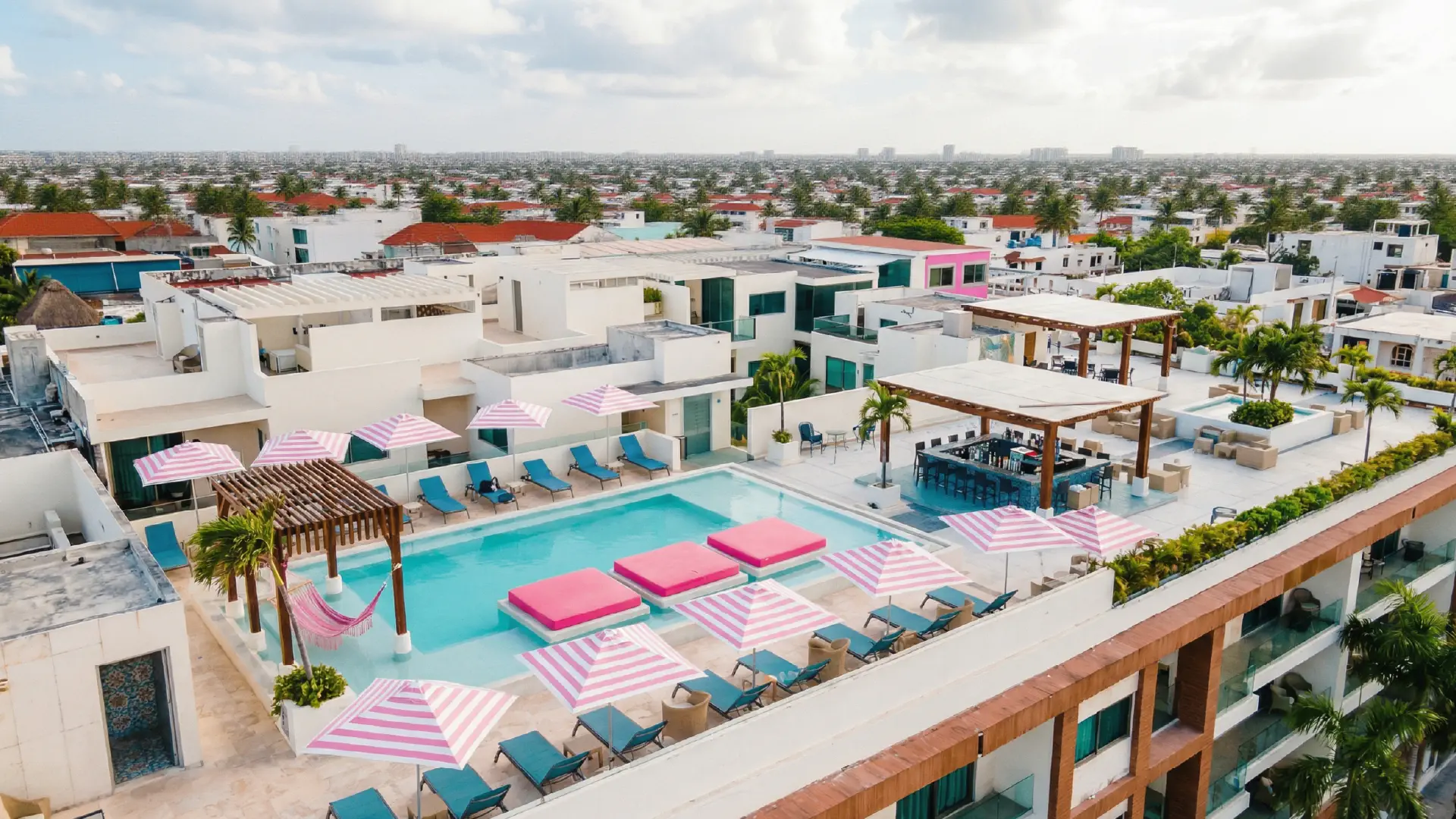 Aerial view of rooftop pool with bar, lounge chairs, and pink striped umbrellas under the sun.