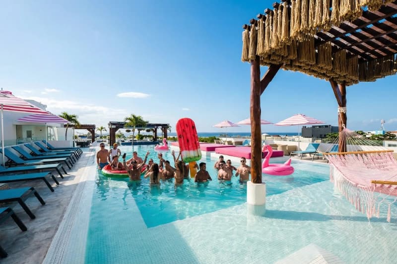 Group of people celebrating in a pool with fun inflatables and striped umbrellas.